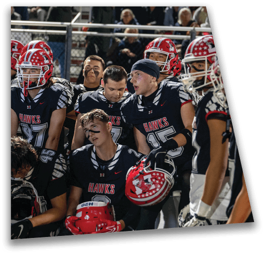 A group of adults wearing black football uniforms with red helmets and face paint are gathered closely together on a sports field with spectators in the background. Description generated by AI