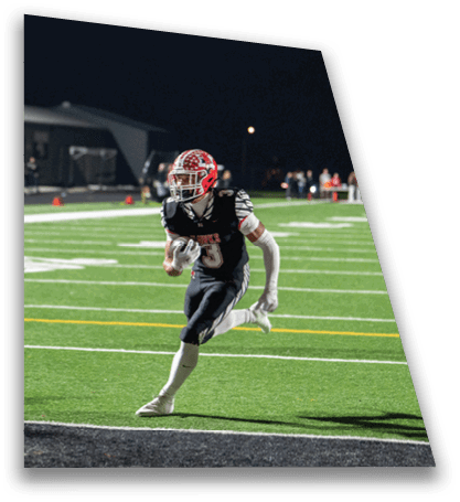 A person wearing a red helmet and black sports uniform is running on a football field at night while holding a football. Description generated by AI