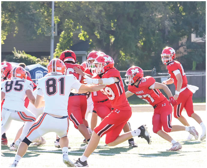 Several people wearing red and white football uniforms are actively engaged in a play on a sunlit outdoor field with trees and a fence in the background. Description generated by AI