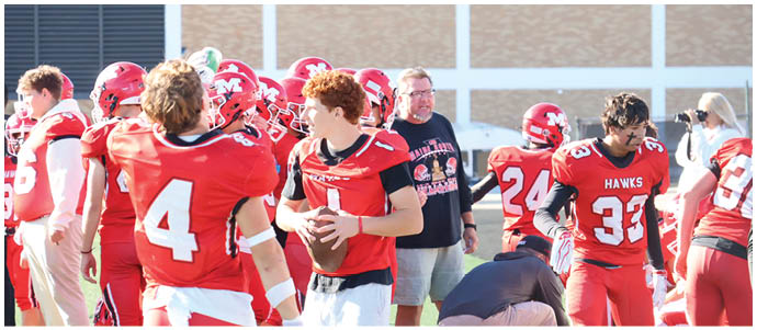 People in red football uniforms are gathered on a field, with one person holding a football and others standing or crouching nearby. Description generated by AI