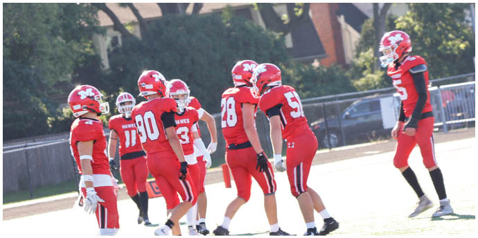 A group of people wearing red football uniforms are gathered on a field while two people in white uniforms stand nearby. Description generated by AI