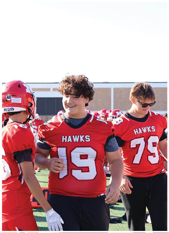 Two people wearing red football jerseys with the word “HAWKS" are interacting on a sports field, with one person smiling and the other wearing a helmet. Description generated by AI