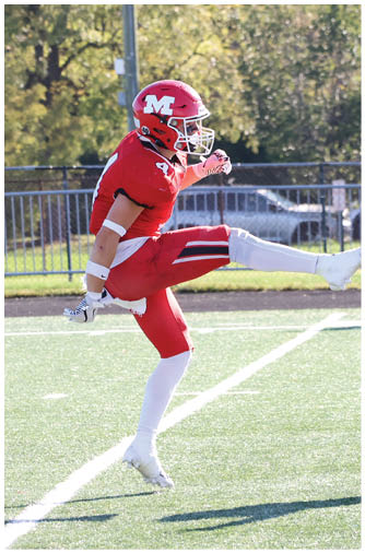 A person in a red football uniform and helmet is in mid-action on a football field with a background of trees and metal barriers. Description generated by AI