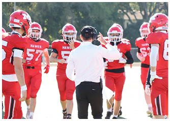 People wearing red football uniforms with the word “HAWKS" are standing and walking outdoors near a person in a white shirt and black pants. Description generated by AI