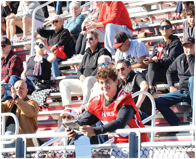 A group of people wearing casual and sports clothing are sitting and standing on bleachers, with one person in a red sports jersey smiling and holding a phone in the foreground. Description generated by AI