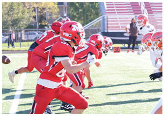 Two opposing football teams in red and white uniforms are lined up facing each other on a sunlit field, preparing for a play. Description generated by AI