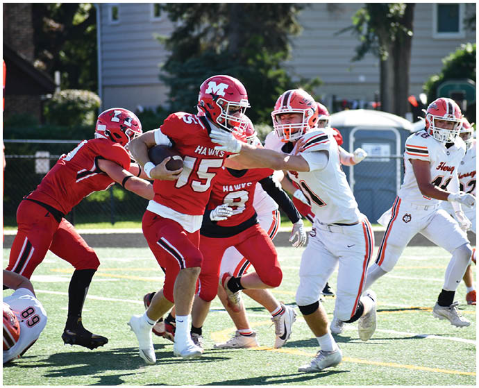 Adults in red and white football uniforms are actively engaged in a game on a sunny outdoor field with one player in red holding the ball while others attempt to block or tackle. Description generated by AI