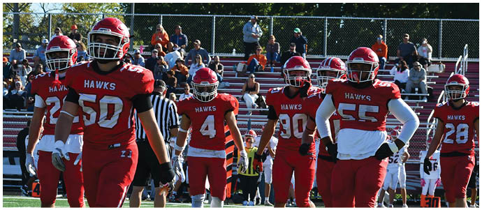 Five people in red football uniforms with “HAWKS" on their jerseys are walking on a football field with spectators in the background. Description generated by AI