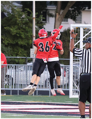 A person wearing sunglasses and a red shirt is leaning on a metal fence near a sports field while another person in a red and black football uniform with the number 36 is jumping in the air. Description generated by AI