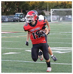 A person wearing a red and black football uniform with the number 19 and the words Maine South is running on a football field while holding a football and being pursued by another player. Description generated by AI