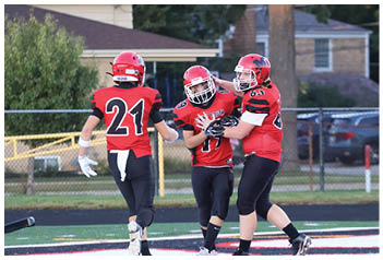 Three people wearing red football uniforms and helmets are on a field, with two of them embracing and the third running toward them. Description generated by AI