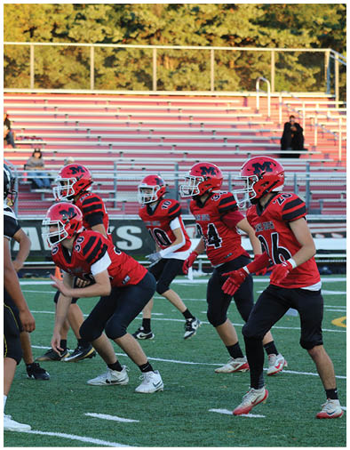 People in red and black football uniforms are lined up on a field facing a person in a white and black uniform during a game. Description generated by AI