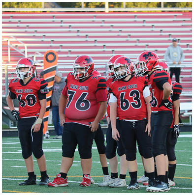 A group of people wearing red and black football uniforms with helmets stand together on a football field near the sidelines. Description generated by AI