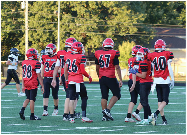 Several people wearing red and black football uniforms are standing on a green field with white yard lines, while one person in a black and white uniform walks in the background. Description generated by AI