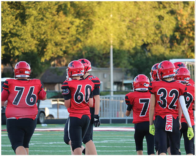 A group of people wearing red and black football uniforms with helmets are standing on a green field with trees and a building in the background. Description generated by AI