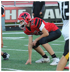 A person in a red football uniform is crouched in a ready position on a football field while another person in a white jersey with the number 12 stands nearby. Description generated by AI