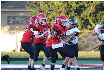 Adults wearing football helmets and uniforms are engaged in a game on a field with one player in red running with the ball while players in white attempt to tackle. Description generated by AI