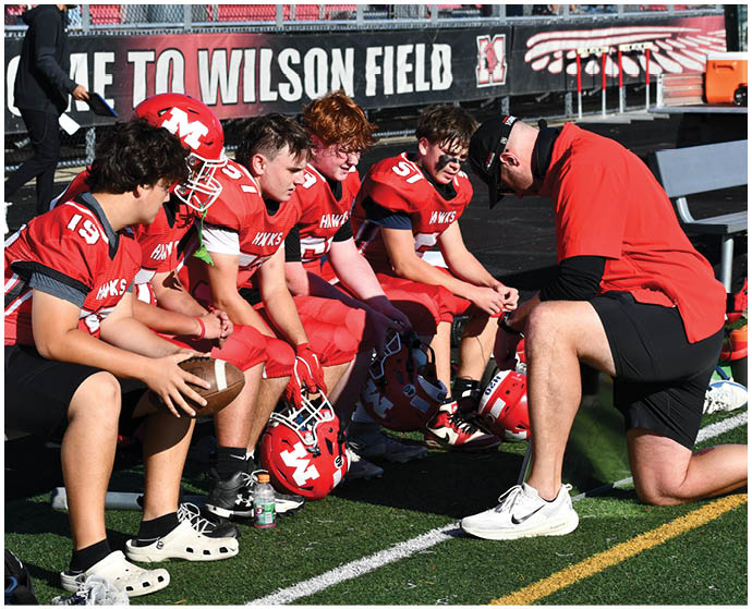 Four people in red football uniforms sit on a bench while another person in a red shirt and black shorts kneels in front of them on a sports field near a sign that reads “WELCOME TO WILSON FIELD." Description generated by AI