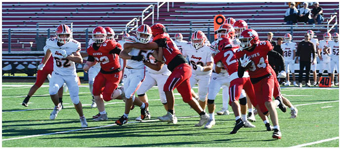 People wearing football uniforms are actively playing on a green field with bleachers and other players visible in the background. Description generated by AI