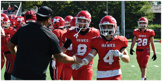 People in red football uniforms with “HAWKS" on the jerseys are on a field, with one person in black handing a water bottle to a player. Description generated by AI