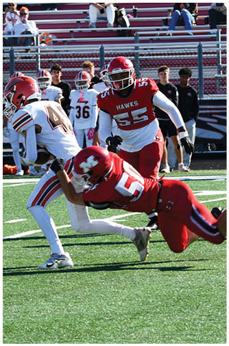 A football player in a red uniform is diving to tackle a player in a white uniform holding the ball on a green field with spectators in the background. Description generated by AI