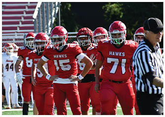 People wearing red football uniforms with “HAWKS" on the jerseys stand on a field while a referee in a black and white striped shirt is nearby. Description generated by AI
