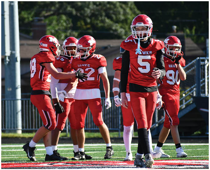 Five people wearing red football uniforms and helmets stand on a football field with one person walking toward the camera and the others interacting behind. Description generated by AI