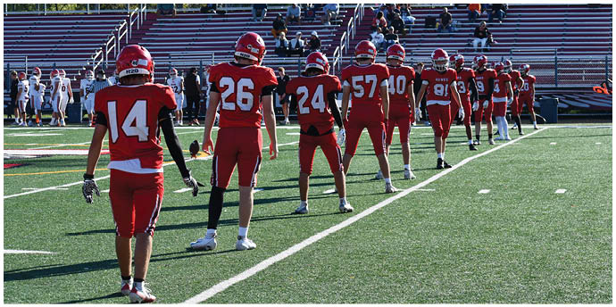 A line of people wearing red football uniforms and helmets stand spaced apart on a green field facing away from the camera with empty bleachers and other players in white uniforms in the background. Description generated by AI