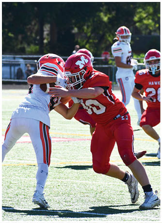 Two people in football uniforms are engaged in a tackle on a sunlit field with other players visible in the background. Description generated by AI