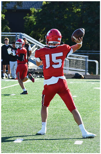 A person wearing a red football helmet and jersey is preparing to throw a football outdoors with trees and a building in the background. Description generated by AI