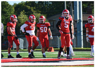 Five people wearing red football uniforms with helmets stand on a football field near the sideline during a game. Description generated by AI