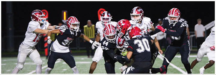 Several people wearing football helmets and uniforms are actively engaged in a play on a football field at night. Description generated by AI