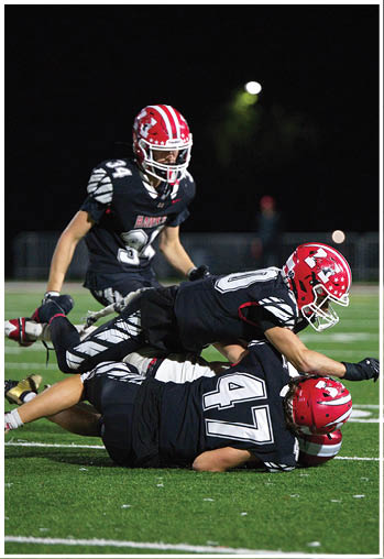 Three football players in black uniforms with red helmets are on a field at night, with two players on the ground and one standing nearby. Description generated by AI