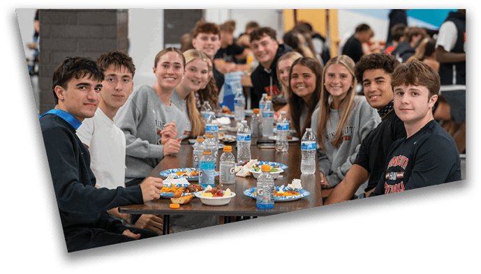 A group of people sitting closely together at a long table in a cafeteria, smiling and eating a meal with water bottles and plates of food in front of them. Description generated by AI