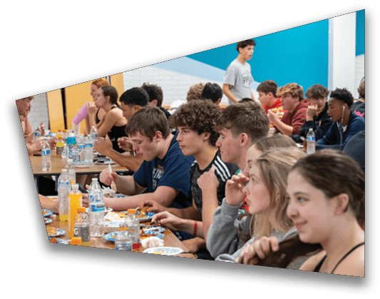 A group of people sitting closely together at long tables eating and drinking in a brightly lit indoor setting. Description generated by AI