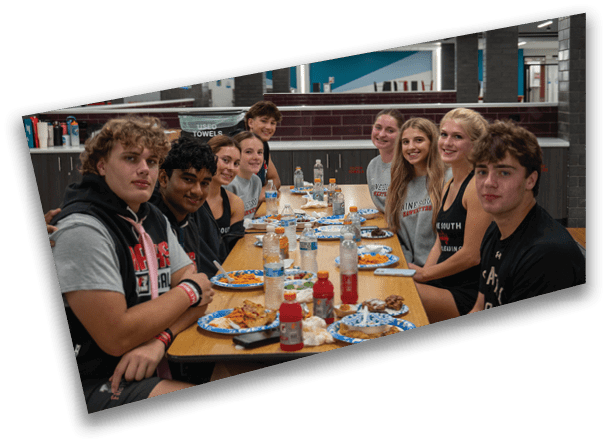 A group of people sitting around a long table in a cafeteria setting, smiling and eating a meal together. Description generated by AI
