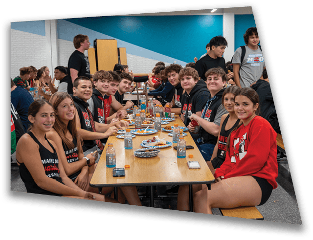 A group of people wearing matching athletic clothing are sitting and standing around a long table in a cafeteria, smiling and engaging with each other. Description generated by AI
