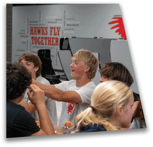 A group of people sitting around a table engaged in conversation with a sign in the background that reads “HAWKS FLY TOGETHER." Description generated by AI