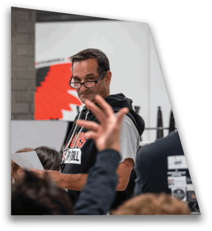 An adult wearing glasses and a hoodie is holding a piece of paper and appears to be speaking to a group of children in a classroom setting. Description generated by AI
