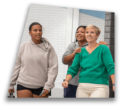 Two adults are smiling and interacting indoors near a black brick wall and a large trash bin, with one person standing behind the other and placing hands on their shoulders. Description generated by AI