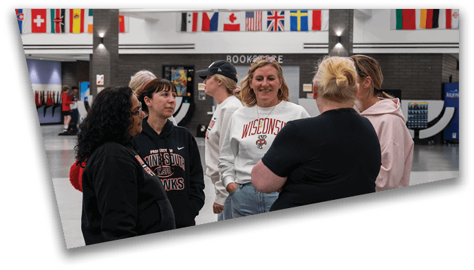 A group of six people wearing casual clothing including sweatshirts with university logos are standing and talking in an indoor space decorated with international flags. Description generated by AI