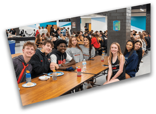 A group of people wearing casual clothing are sitting and smiling around a wooden table in a busy cafeteria setting. Description generated by AI