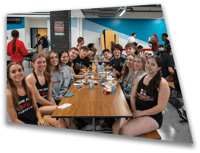 A group of people wearing matching casual sportswear sit closely together at a long cafeteria table with food and drinks, smiling and engaging in a social setting. Description generated by AI