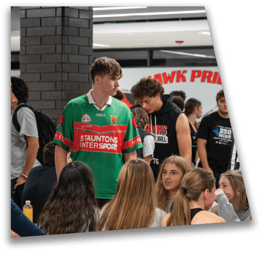 A group of people are gathered indoors with one person standing in a green sports jersey while others sit or stand nearby engaged in conversation. Description generated by AI
