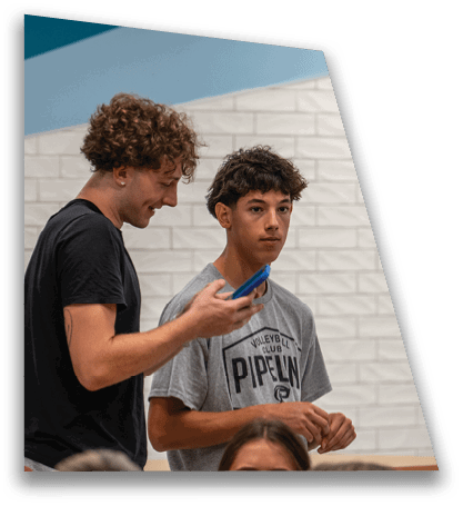 Two people with curly hair stand indoors near a white tiled wall, one wearing a black shirt looking at a phone and the other wearing a gray volleyball club t-shirt looking forward. Description generated by AI