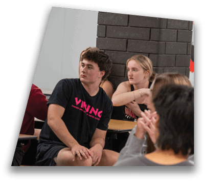 A group of people sitting at desks in a classroom setting, with two individuals in the center wearing black shirts with pink text and appearing attentive. Description generated by AI