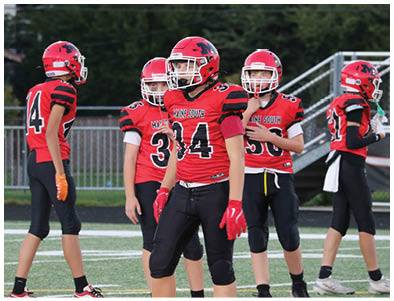 Five people wearing red and black football uniforms stand on a field with a fence and trees in the background. Description generated by AI