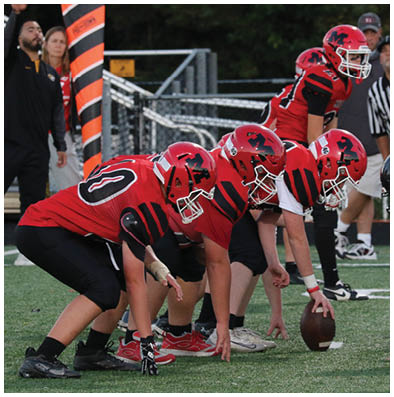 Four people in red and black football uniforms are lined up on a field with one holding a football ready to snap it. Description generated by AI
