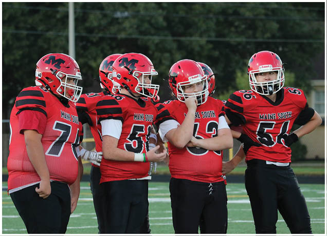 Four people wearing red and black football uniforms and helmets stand together on a football field, appearing to be in conversation or planning. Description generated by AI