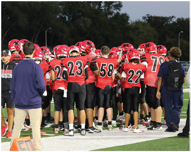 A group of people wearing red football uniforms and helmets are gathered on a field while two people in casual clothing stand nearby. Description generated by AI